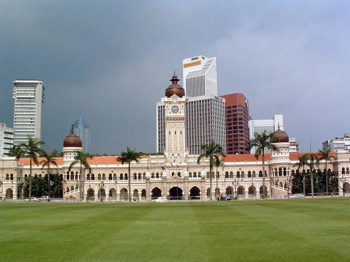 Edifício Sultan Abdul Samad e sua torre do relógio, palmeiras e prédios de Kuala Lumpur vistos do gramado da praça Merdeka.