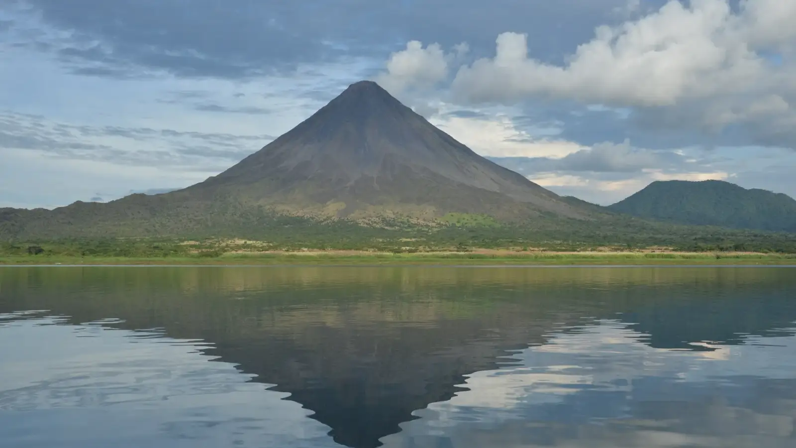 O cone simétrico do vulcão Arenal refletido na água calma do lago Arenal, sob nuvens, com floresta na margem.