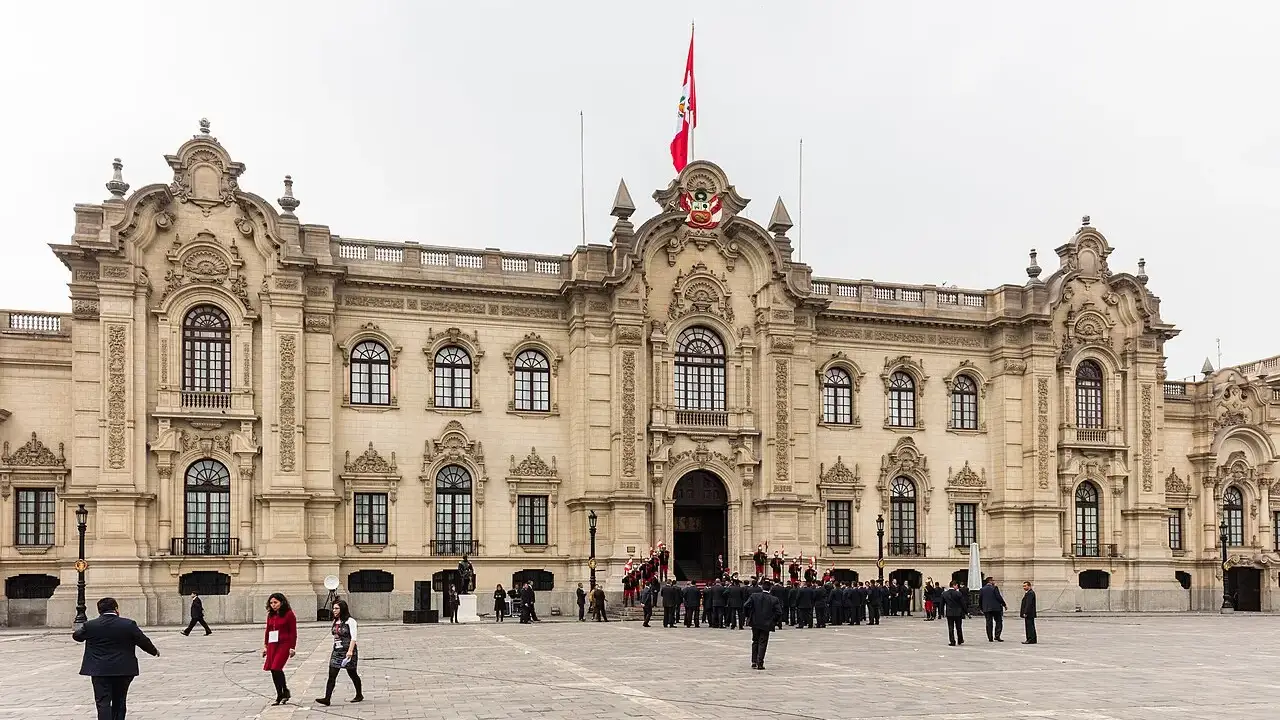 Fachada do Palacio de Gobierno na praça, com a bandeira do Peru e pessoas no piso de pedra.