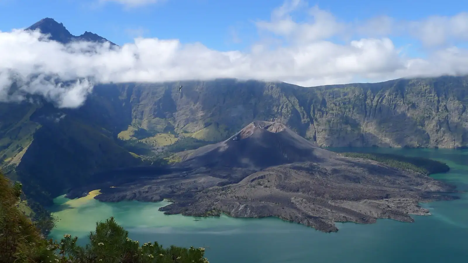 La caldeira du Gunung Rinjani, à Lombok : le lac Segara Anak, aux eaux bleu vert, autour du cône sombre du Gunung Barujari, sous la paroi du cratère, avec des nuages bas et le sommet du Rinjani en haut à gauche.