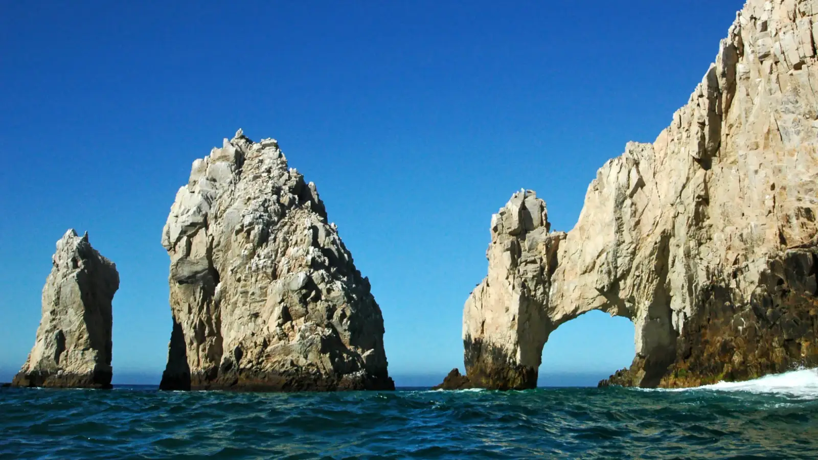 O Arco de Cabo San Lucas e duas agulhas de rocha clara saindo do mar azul em Land's End, sob céu sem nuvens.