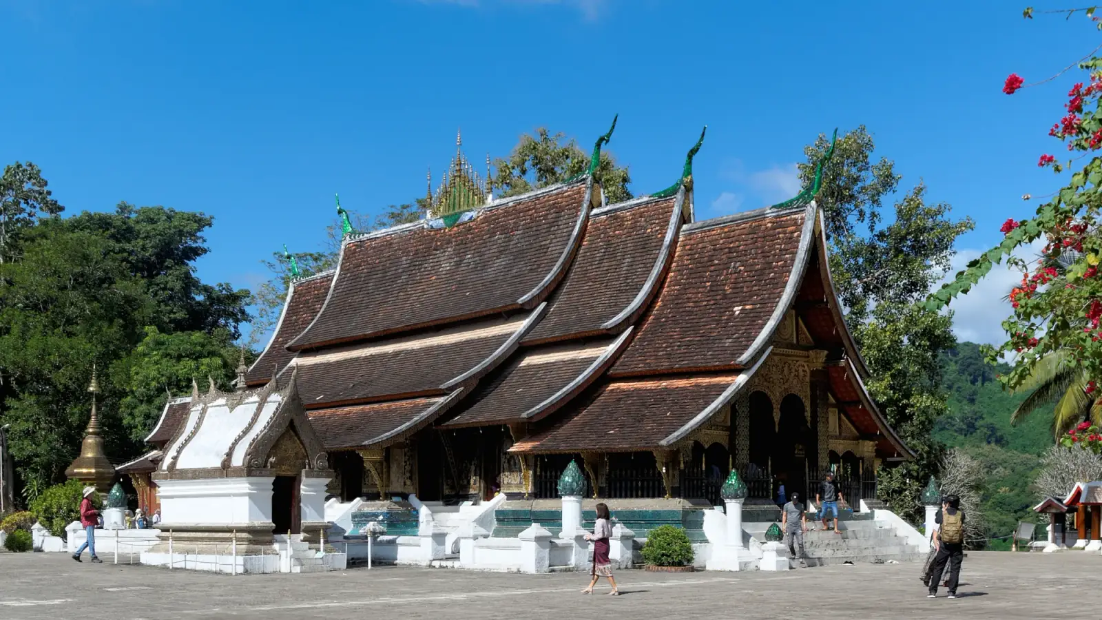 O sim do Wat Xieng Thong, em Luang Prabang, com os telhados escuros sobrepostos que descem quase até o chão, uma capela branca ao lado, o pátio de pedra e árvores sob o céu azul.