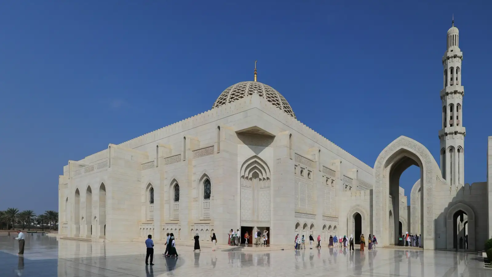 The prayer hall of the Sultan Qaboos Grand Mosque in white marble, with its latticed dome, a minaret and an arched portal, and visitors on the polished marble courtyard under a blue sky.