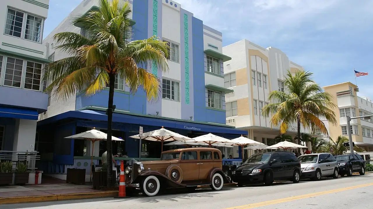 Prédios Art Déco pintados de azul e branco na Ocean Drive, com palmeiras, um carro antigo estacionado e mesas com guarda-sóis na calçada, sob céu azul.