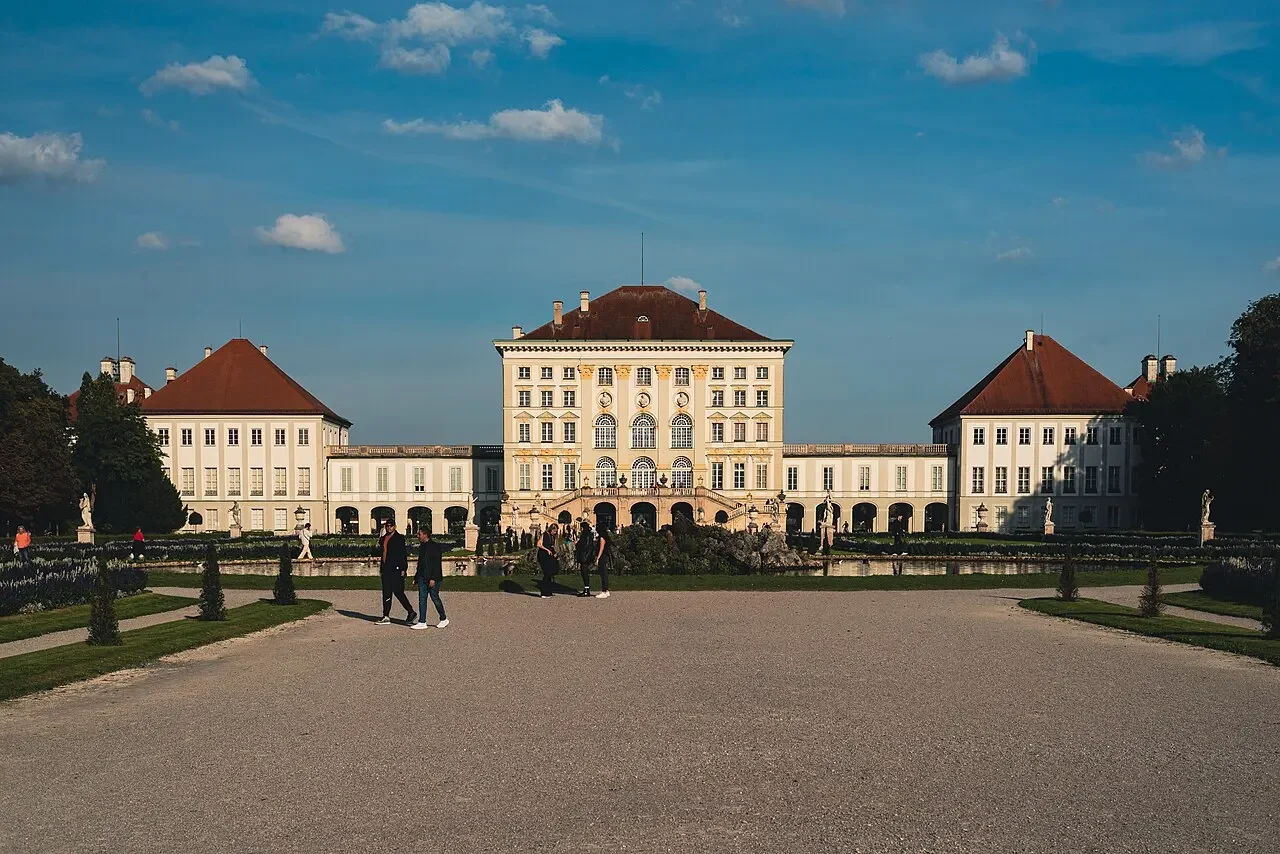 Palácio de Nymphenburg em Munique, com pavilhão central, alas laterais, jardim, visitantes e caminho frontal de cascalho.