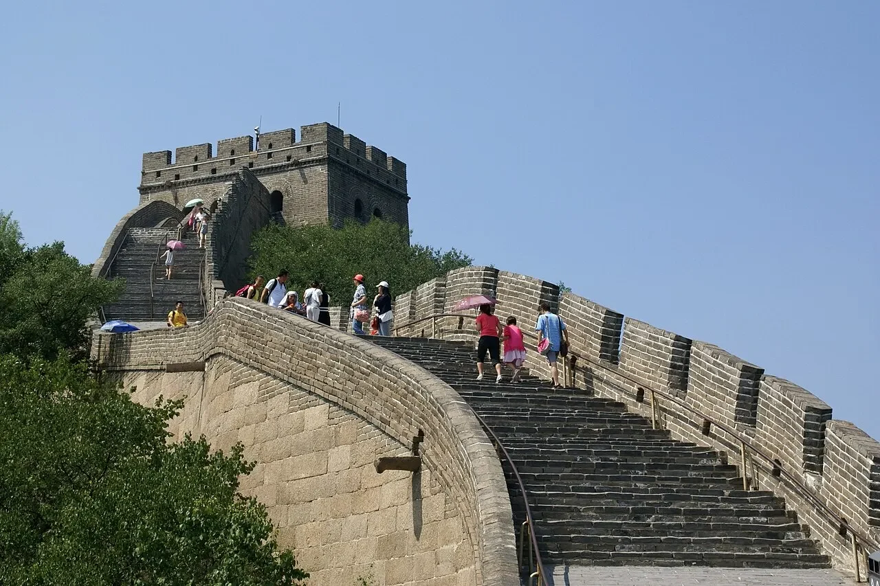 Trecho íngreme da Muralha da China com degraus de pedra subindo até uma torre de vigia, visitantes caminhando com guarda-sóis sob céu azul.
