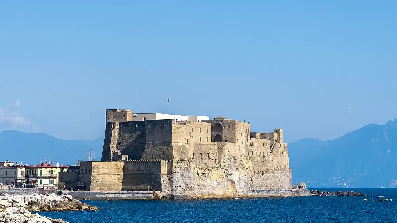 Castel dell'Ovo, em Nápoles, fortaleza de pedra sobre um ilhéu no golfo, com montanhas ao fundo.