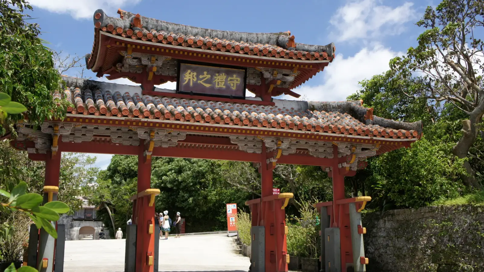 The Shureimon gate of Shuri Castle in Naha, with two red-tiled roofs, the plaque with gold characters and red pillars among trees, under a blue sky