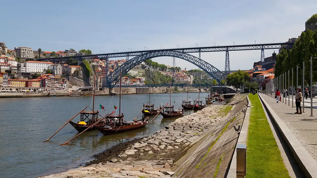 Ponte de ferro Luís I em arco sobre o rio Douro, com barcos rabelos ancorados na margem e casas coloridas subindo a encosta, sob céu azul claro.