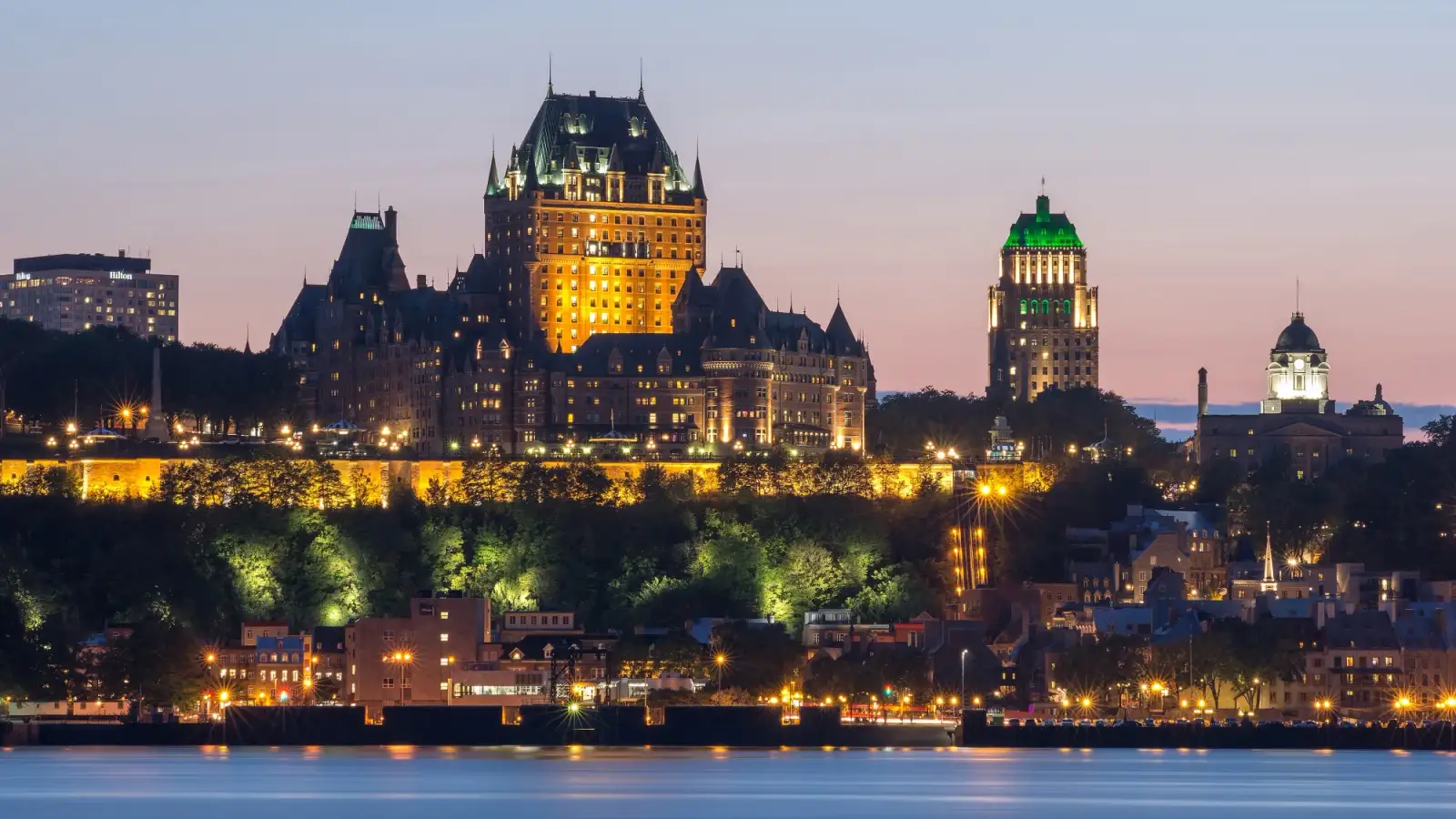O Château Frontenac iluminado no alto do cabo, sobre a cidade velha e o rio São Lourenço, visto de Lévis ao anoitecer.