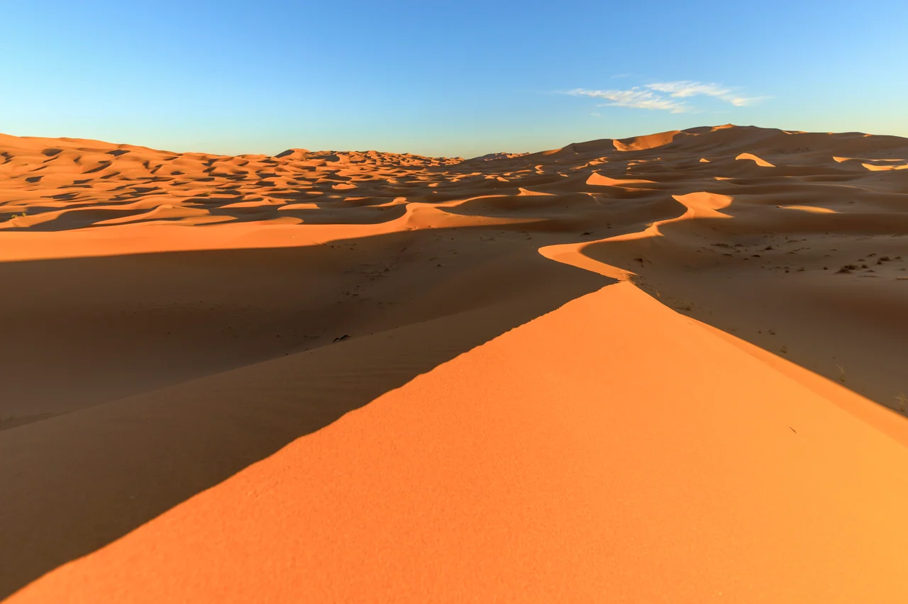 Dunas do Erg Chebbi, na parte marroquina do Saara, com céu azul, curvas iluminadas e sombreadas ao longe e uma duna alaranjada em primeiro plano.
