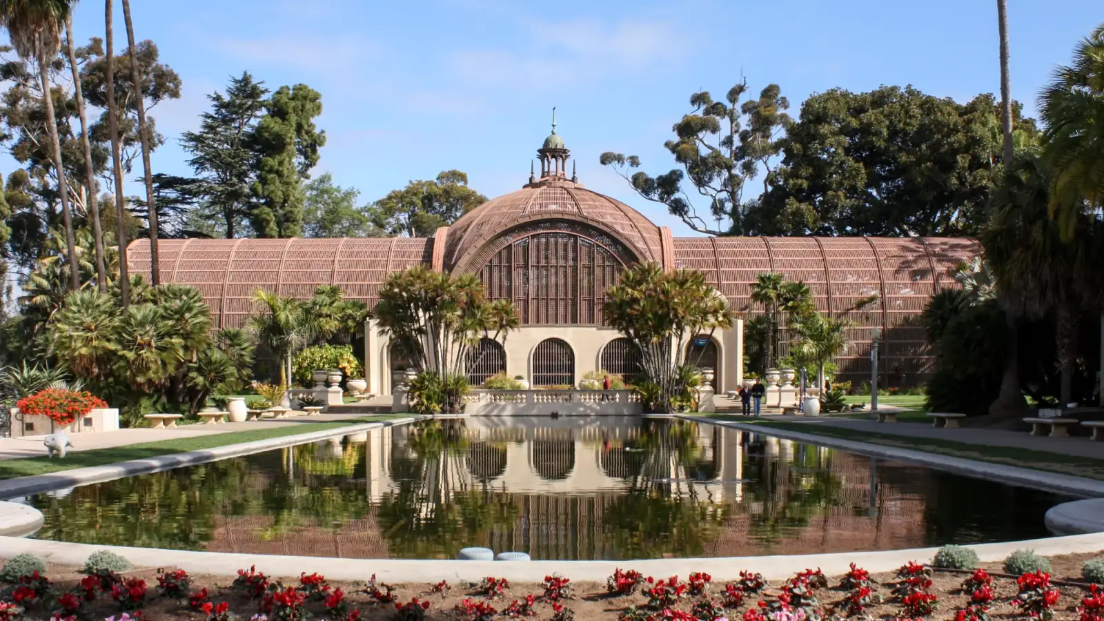 O Botanical Building do Balboa Park, de ripas de madeira com cúpula, refletido no lago das ninfeias, com palmeiras e canteiros floridos à frente.