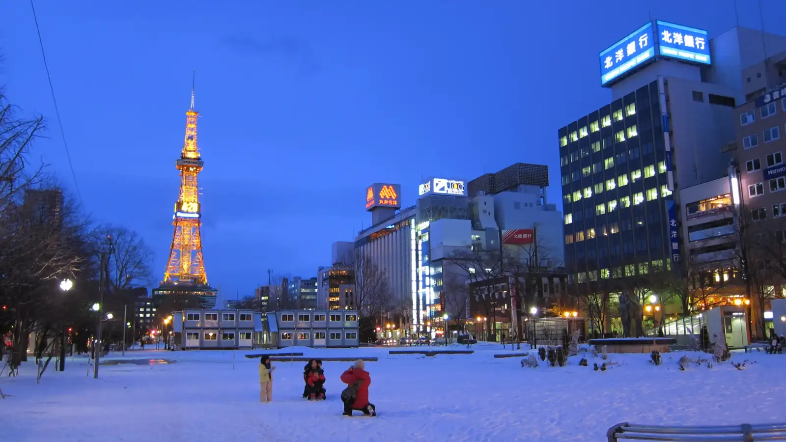 Odori Park covered in snow at dusk, with the Sapporo TV Tower lit in orange on the left, office buildings with lit windows on the right and a few small figures in the snow.