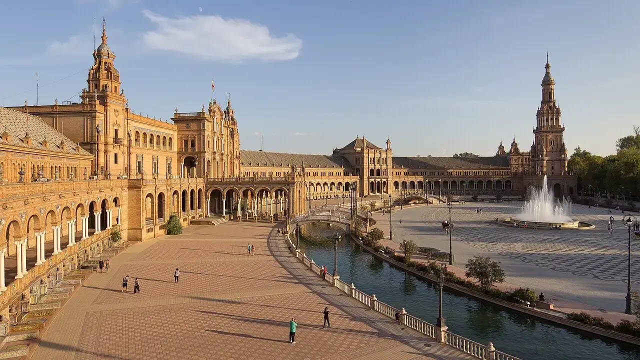 A Plaza de España, com o edifício semicircular de tijolo e torres, o canal e pessoas caminhando na praça.