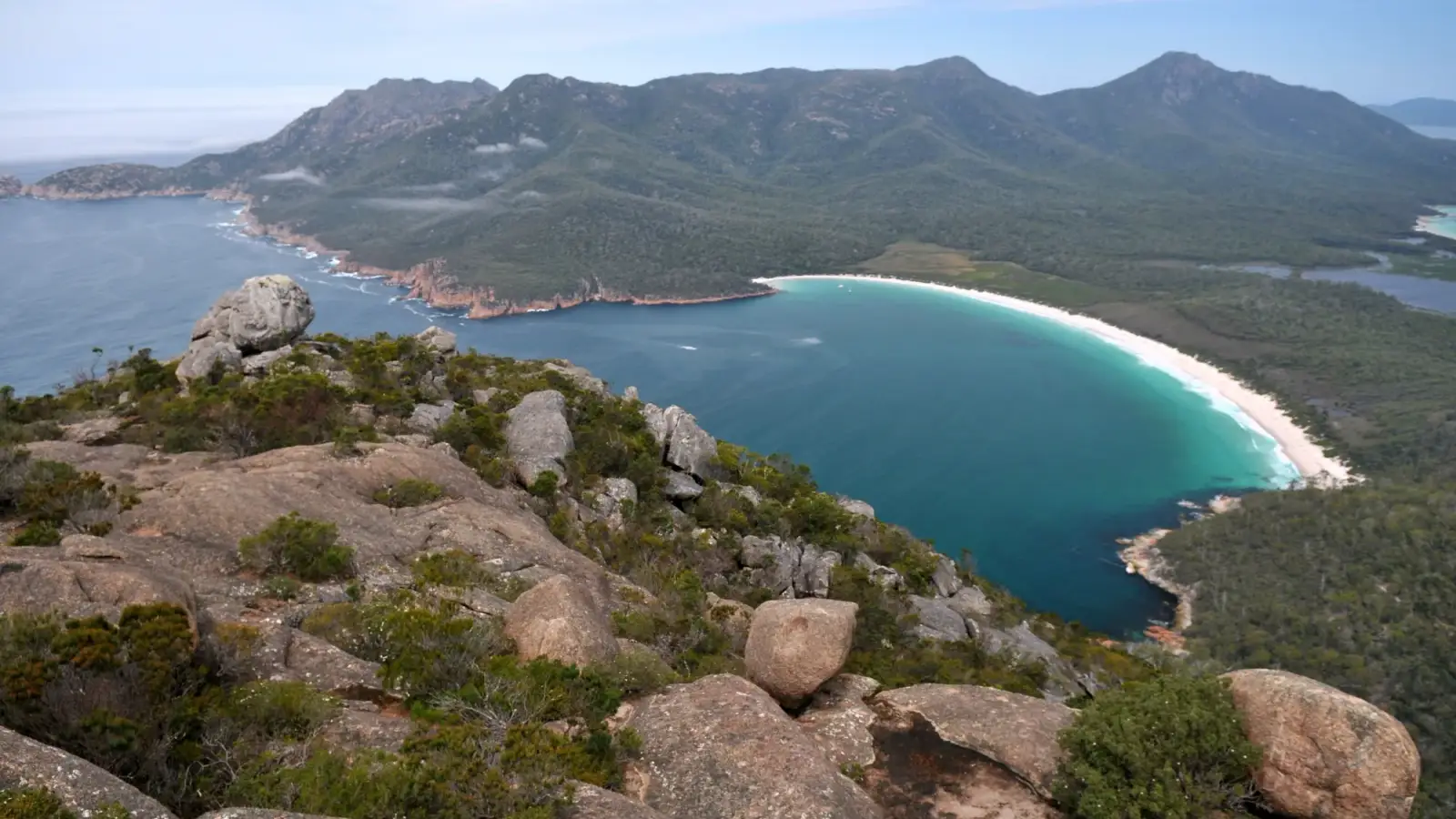 Wineglass Bay, a crescent of turquoise water and white sand, with the forested Hazards mountains behind it, seen from above among granite boulders.