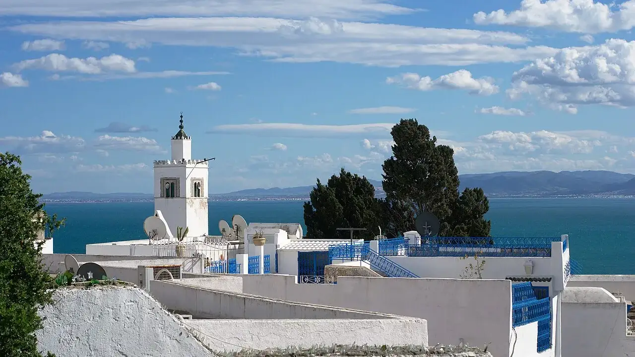 Telhados brancos com grades azuis de Sidi Bou Saïd, um minarete ao centro e o mar Mediterrâneo ao fundo, sob céu com nuvens.