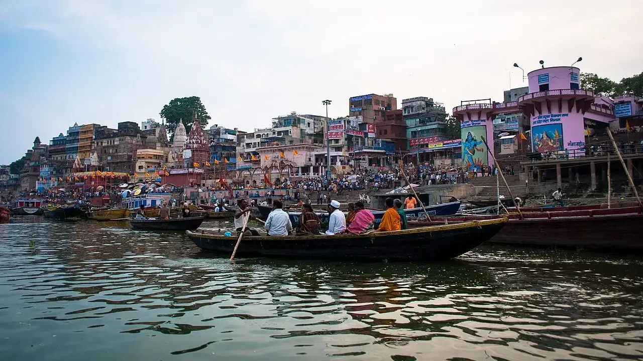 Barcos de madeira lotados de passageiros no rio Ganges em frente ao Dashashwamedh Ghat, com multidão nas escadarias e prédios coloridos ao fundo.