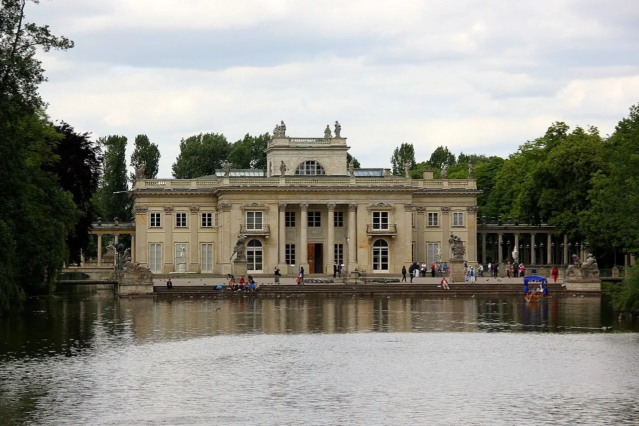 Palácio sobre a Ilha no parque Łazienki, em Varsóvia, com esculturas no telhado, árvores, visitantes e um pequeno barco junto à margem; água e reflexos à frente, céu nublado ao fundo.