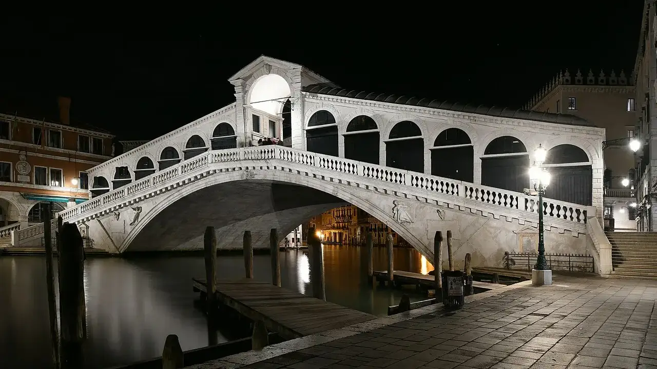 Ponte de Rialto, em Veneza, com o arco de pedra e as lojas em cima, iluminada à noite sobre o Grande Canal.