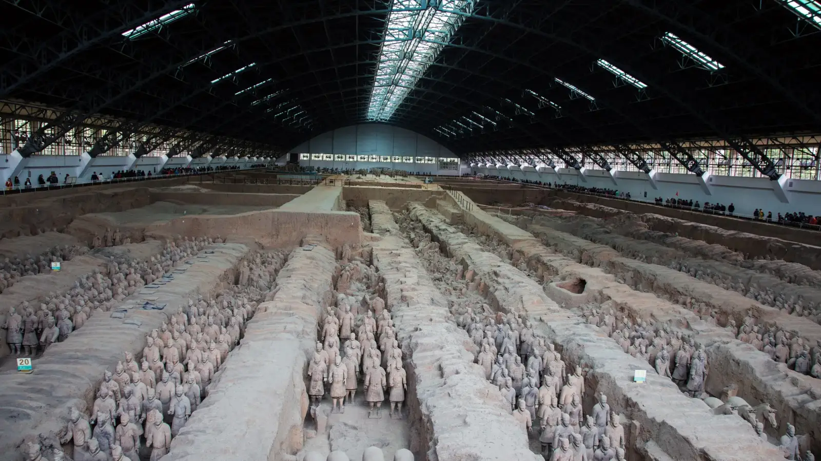 Pit 1 of the Terracotta Army in Xi'an: rows of clay warriors standing in the excavated corridors between earth walls, under the metal roof of the hall, with small visitors on the gallery in the distance.
