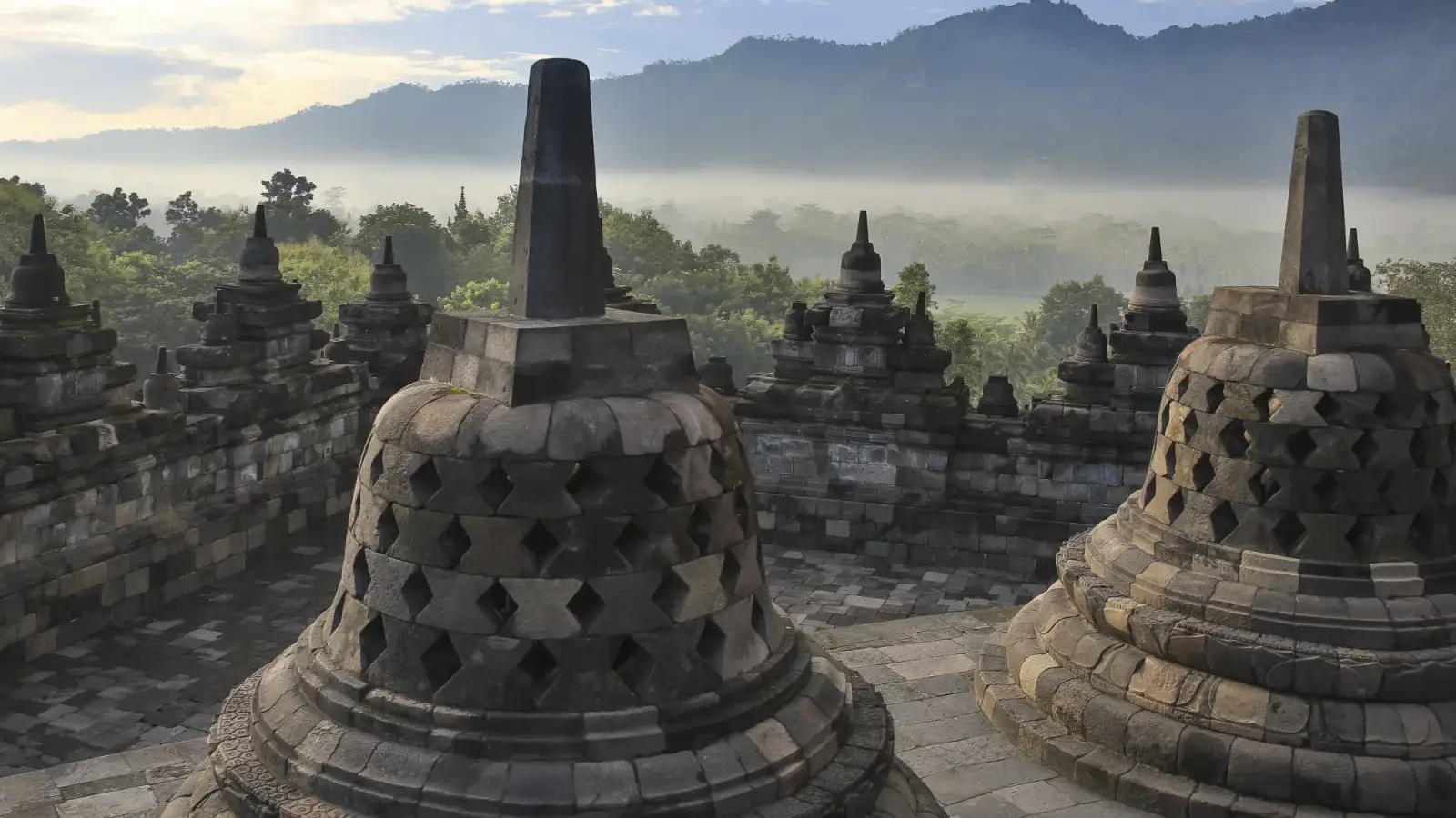 Estupas de pedra perfurada no terraço do Borobudur, com outras estupas menores atrás, mata envolta em névoa e uma serra ao fundo sob o céu claro da manhã.
