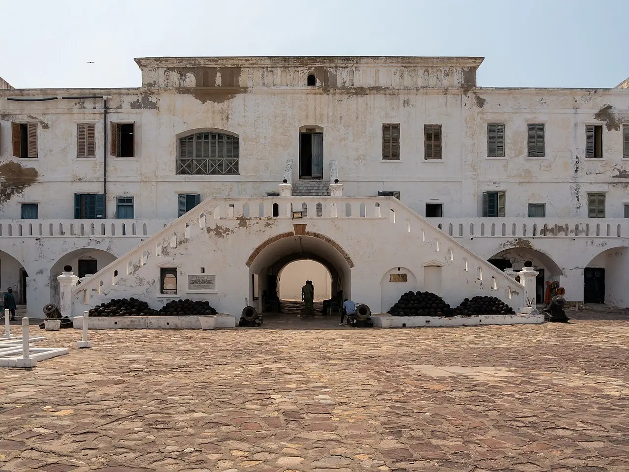 Cour pavée du château de Cape Coast, avec une façade blanche usée, un double escalier au-dessus d’une arche et des piles de boulets de canon.
