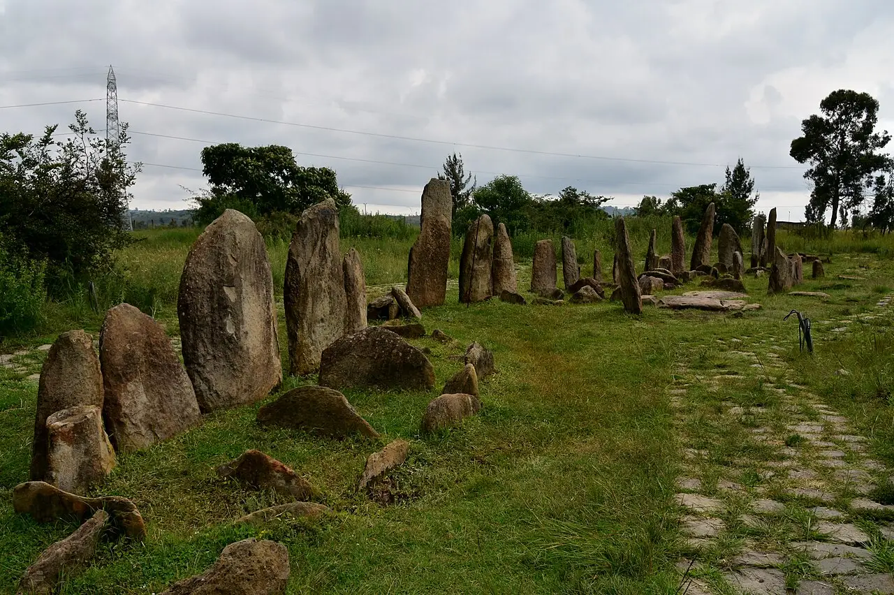 Campo de estelas de pedra de Tiya, com dezenas de pedras erguidas sobre grama, um caminho de lajes à direita e céu nublado.