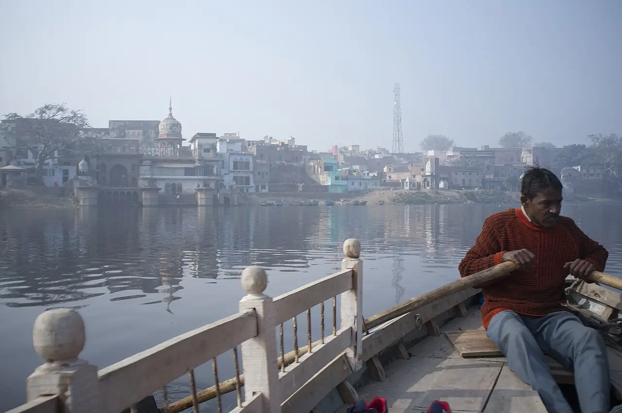 Margem do rio em Mathura vista de um barco a remo, com casas e uma cúpula à beira da água e um remador em primeiro plano.