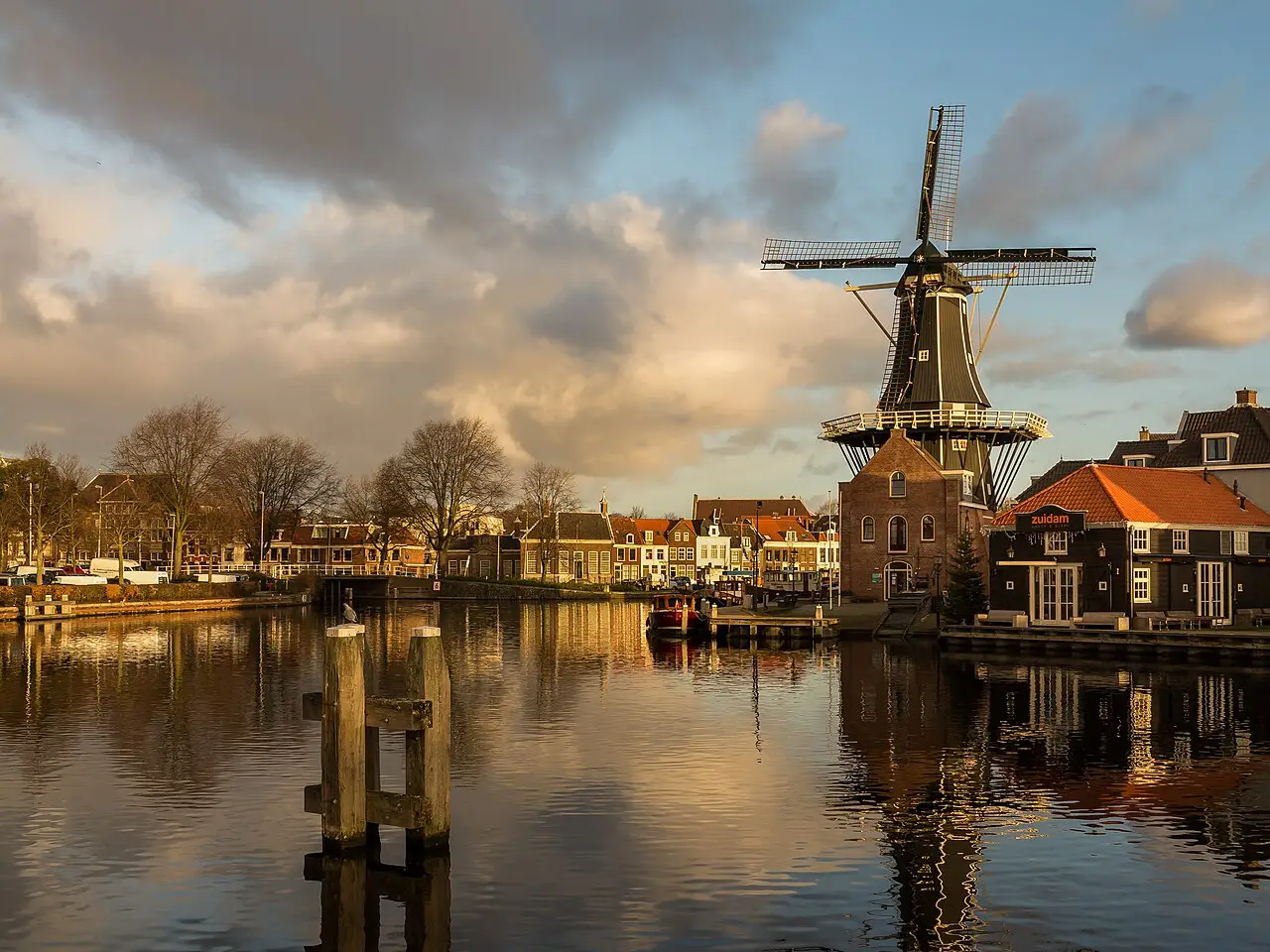Moinho De Adriaan à beira do rio Spaarne em Haarlem, com casas de tijolo ao fundo e o reflexo das nuvens e da luz dourada na água.