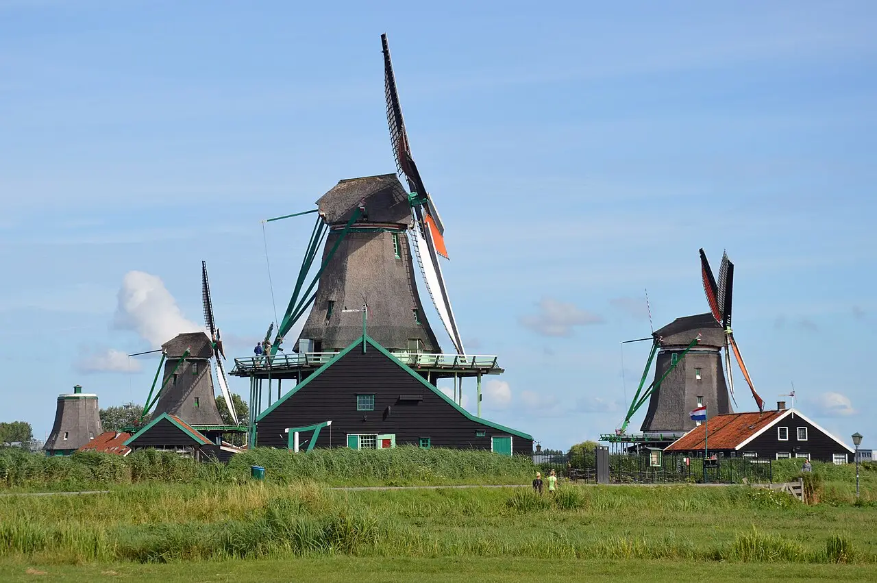 Moinhos de vento de madeira escura em Zaanse Schans, com casas de telhado vermelho, campo de grama e céu azul ao fundo.