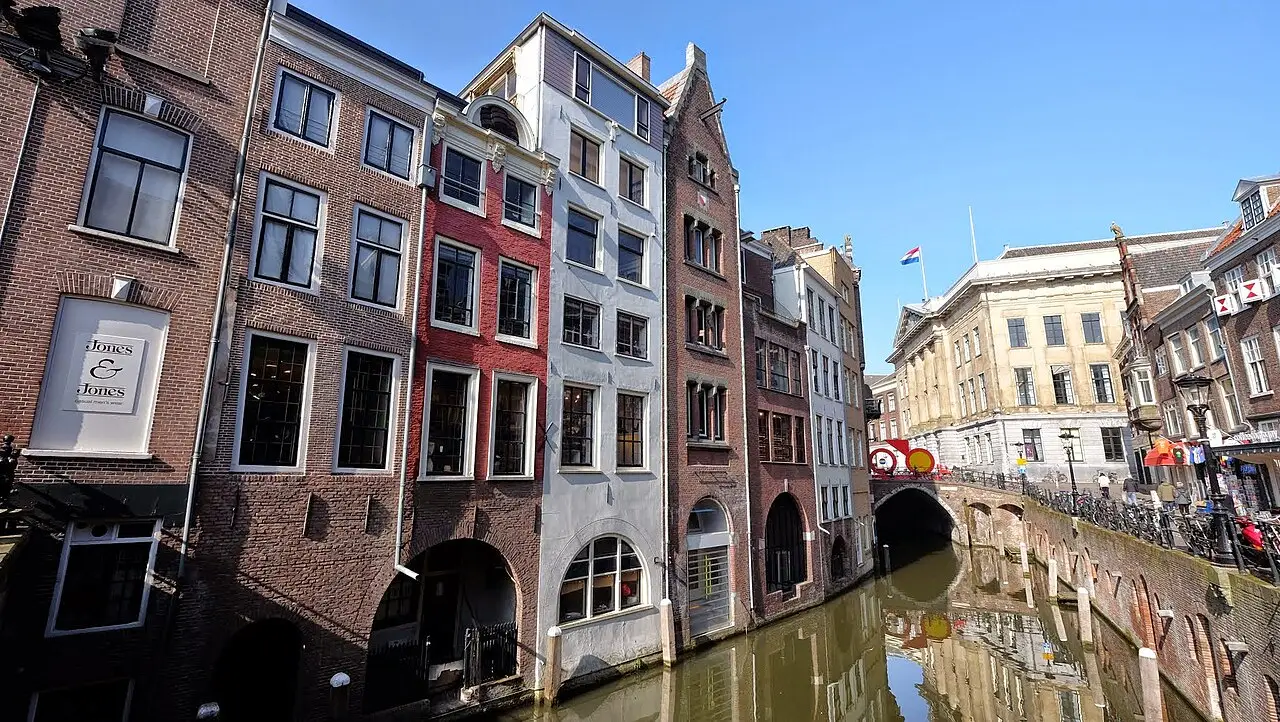 Canal no centro de Utrecht, com fachadas de tijolo estreitas à beira da água, uma ponte de arco ao fundo e bandeira holandesa ao alto.
