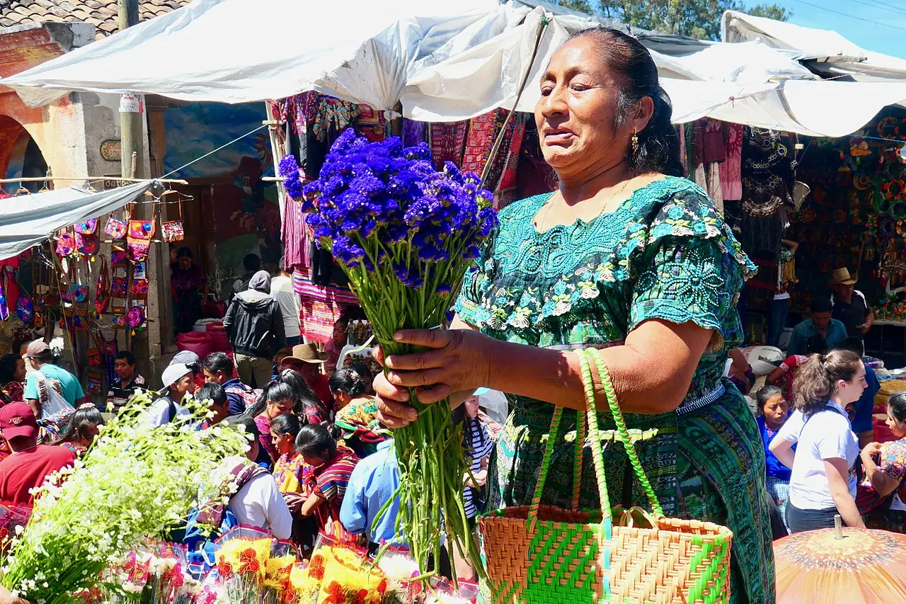 Vendedora com um buquê de flores roxas no mercado de Chichicastenango, com barracas de tecidos coloridos e clientes ao fundo.