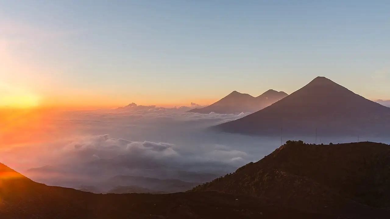 Vista do Vulcão Pacaya com sol baixo no horizonte, um mar de nuvens no vale e os cones de outros vulcões da Guatemala ao fundo.