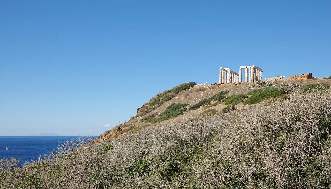 Colunas do Templo de Poseidon no alto de uma colina no Cabo Sunião, com o mar azul à esquerda e vegetação seca em primeiro plano.