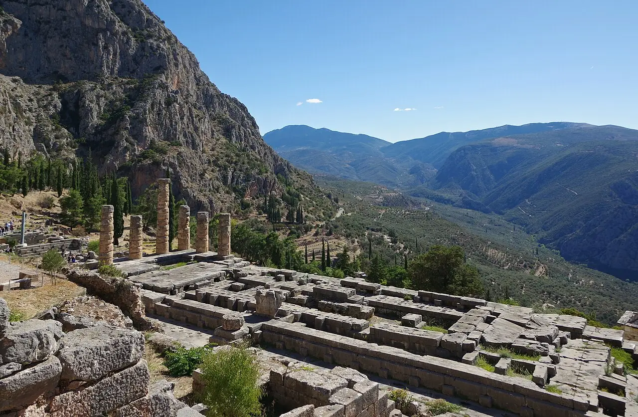 Ruínas do Templo de Apolo em Delfos, com colunas restantes, blocos de pedra do piso e um vale de montanhas ao fundo.