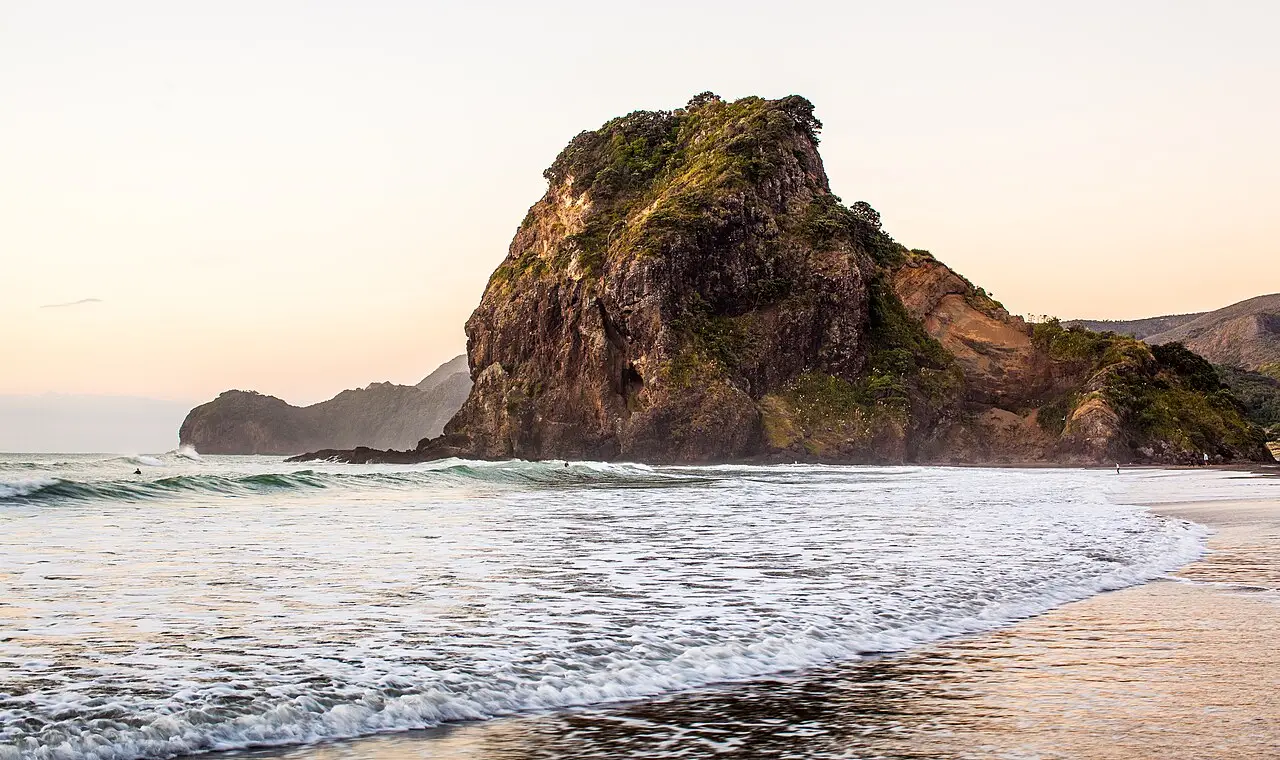 Lion Rock, grande rocha coberta de vegetação na praia de Piha, com ondas chegando à areia molhada sob um céu de tons claros.