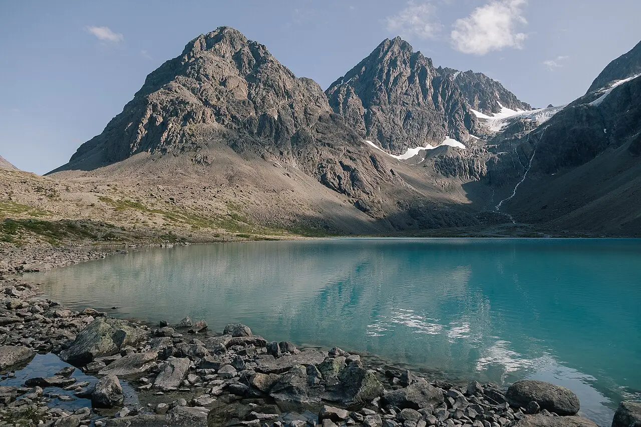 Lago Blåvatnet de água turquesa nos Alpes de Lyngen, com picos rochosos e manchas de neve ao fundo e pedras na margem.