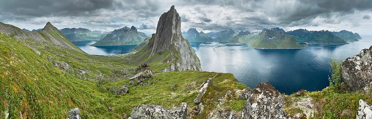 Vista panorâmica de Senja a partir de uma crista, com o pico pontiagudo de Segla, um fiorde escuro e montanhas cobertas de verde.