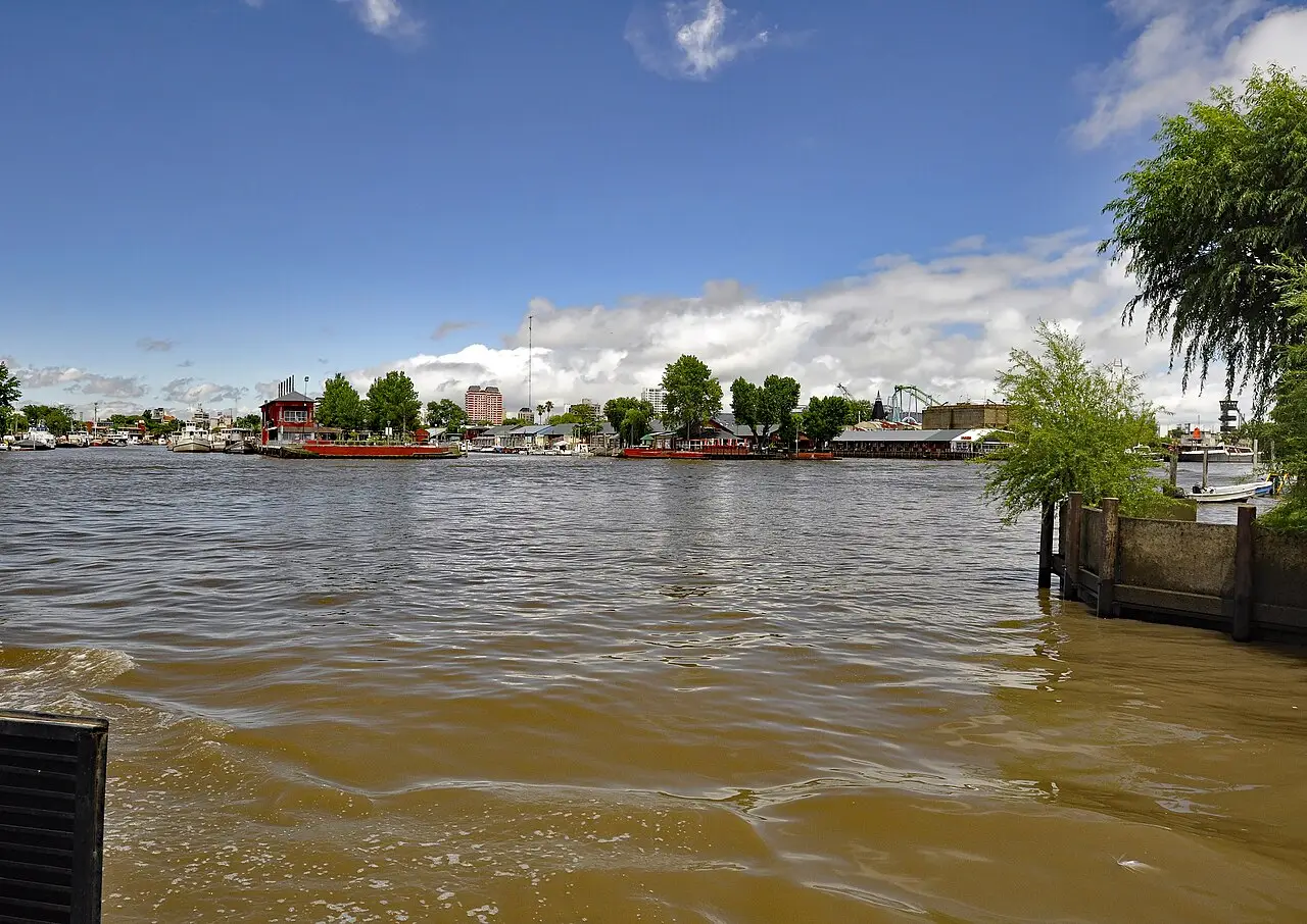 Rio de águas marrons em Tigre, com barcos atracados na margem oposta, árvores, edifícios e brinquedos de um parque ao fundo.