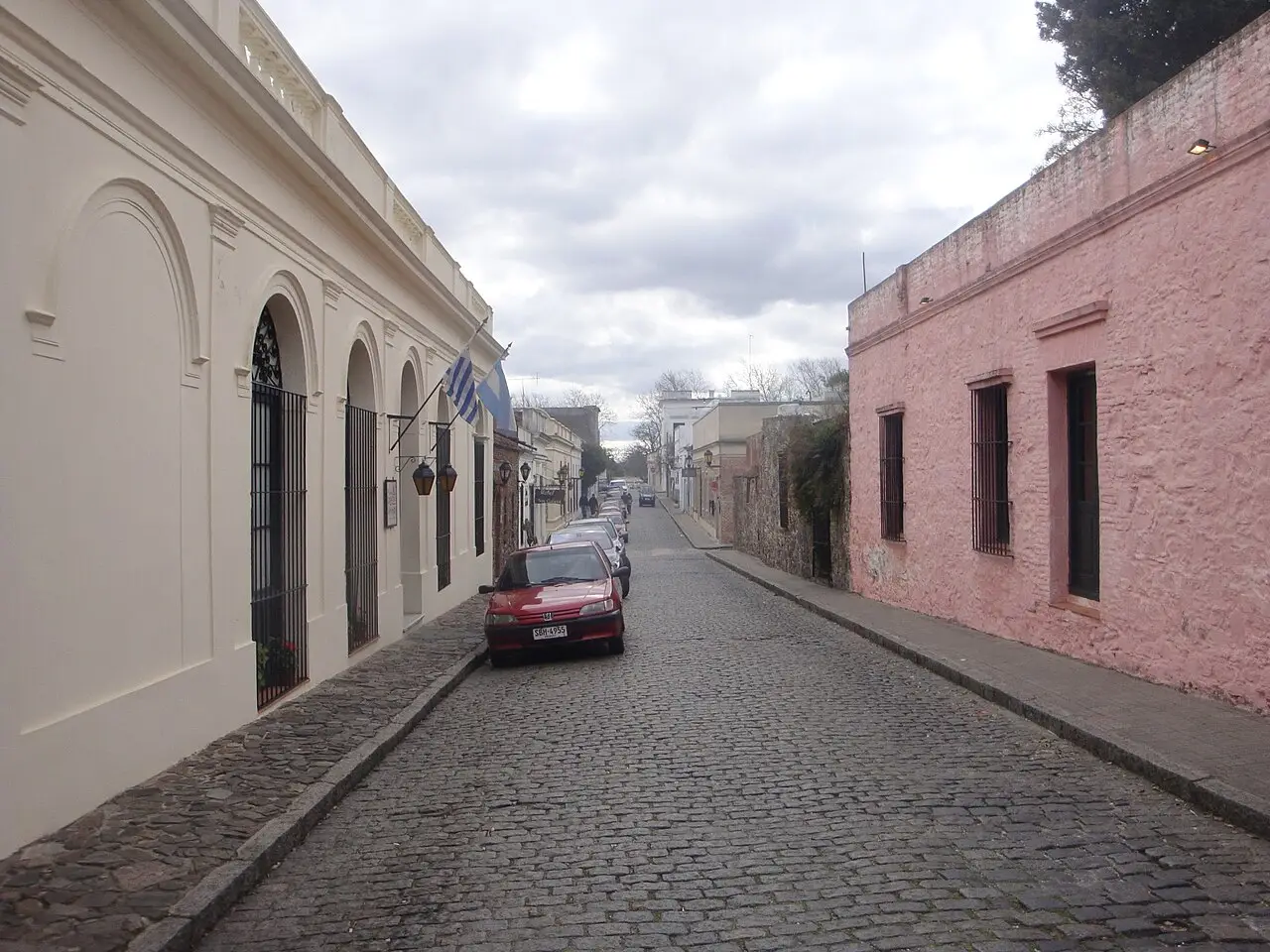 Rua de paralelepípedos em Colonia del Sacramento, com casas baixas de fachada creme e rosa e bandeiras do Uruguai e da Argentina.