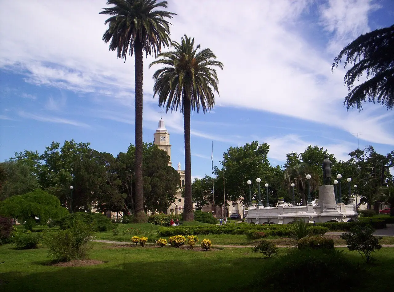 Plaza Ruiz de Arellano em San Antonio de Areco, com palmeiras altas, jardins, um monumento com estátua e uma torre de igreja ao fundo.