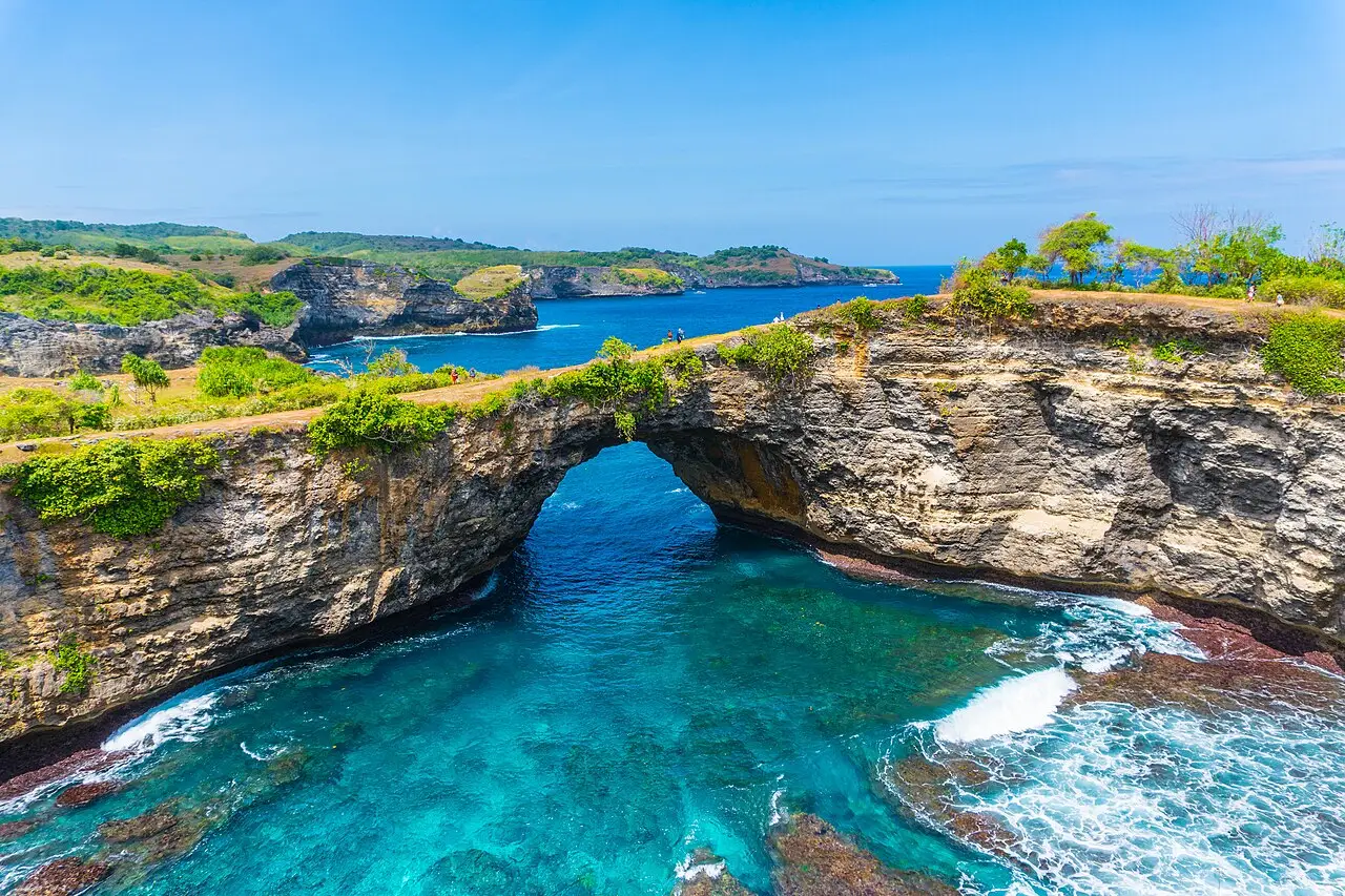 Arco natural de rocha sobre uma enseada de água turquesa em Nusa Penida, com falésias verdes e o mar aberto ao fundo.
