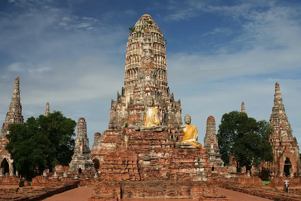Ruínas de tijolo do Wat Chaiwatthanaram em Ayutthaya, com um prang central alto, duas estátuas de Buda com mantos alaranjados e torres menores.