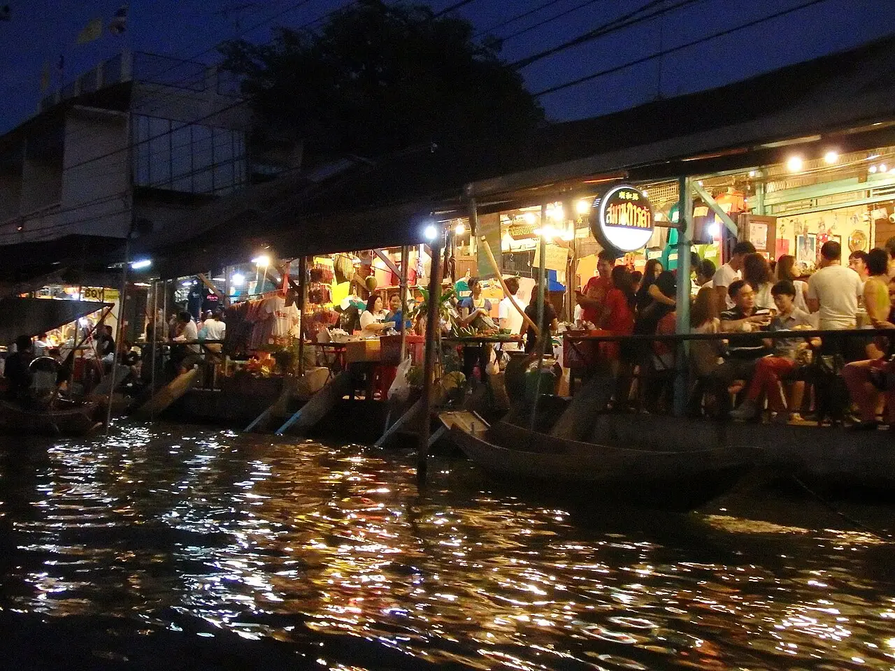 Lojas e restaurantes iluminados à beira do canal em Amphawa à noite, com barcos de madeira na água e gente reunida na margem.