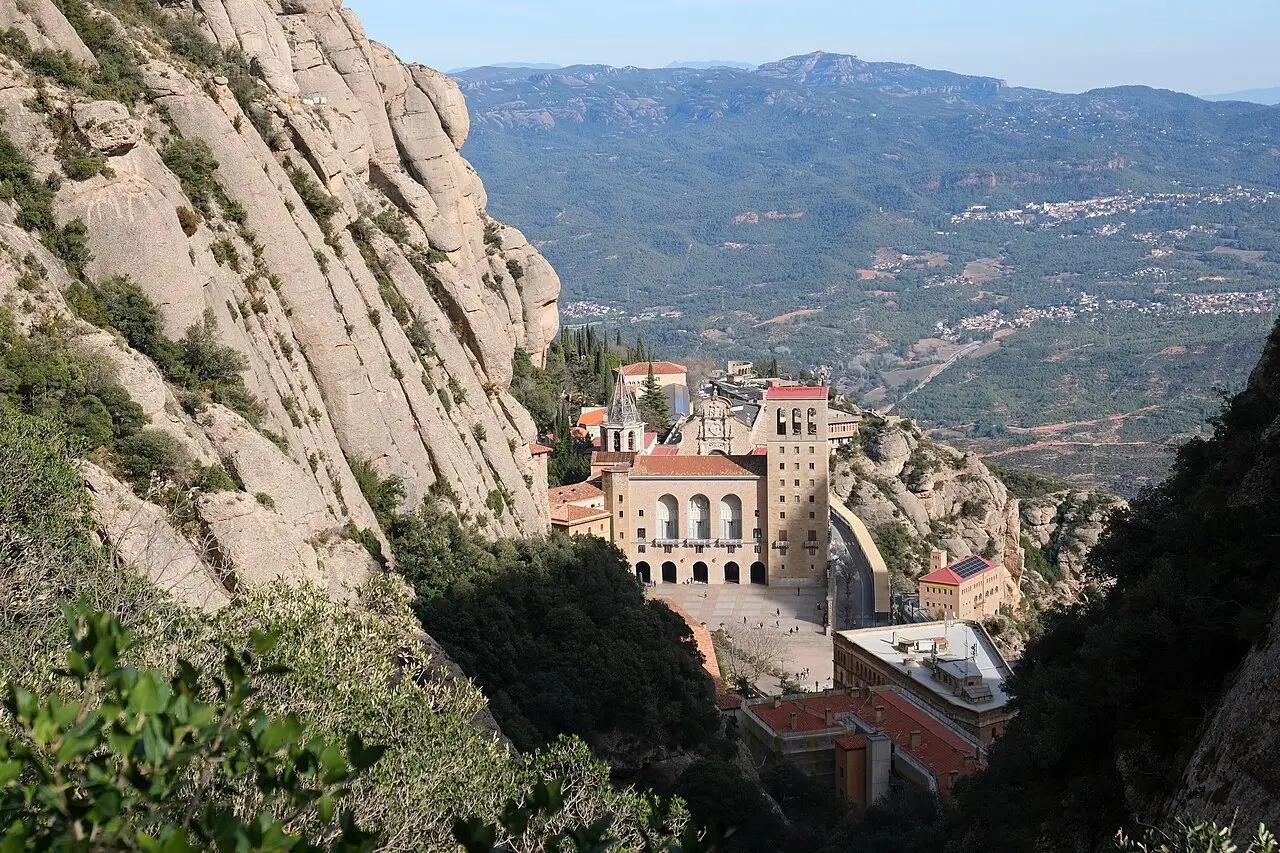 Mosteiro de Montserrat visto do alto, encaixado entre paredões de rocha clara, com um vale de montanhas e povoados ao fundo.