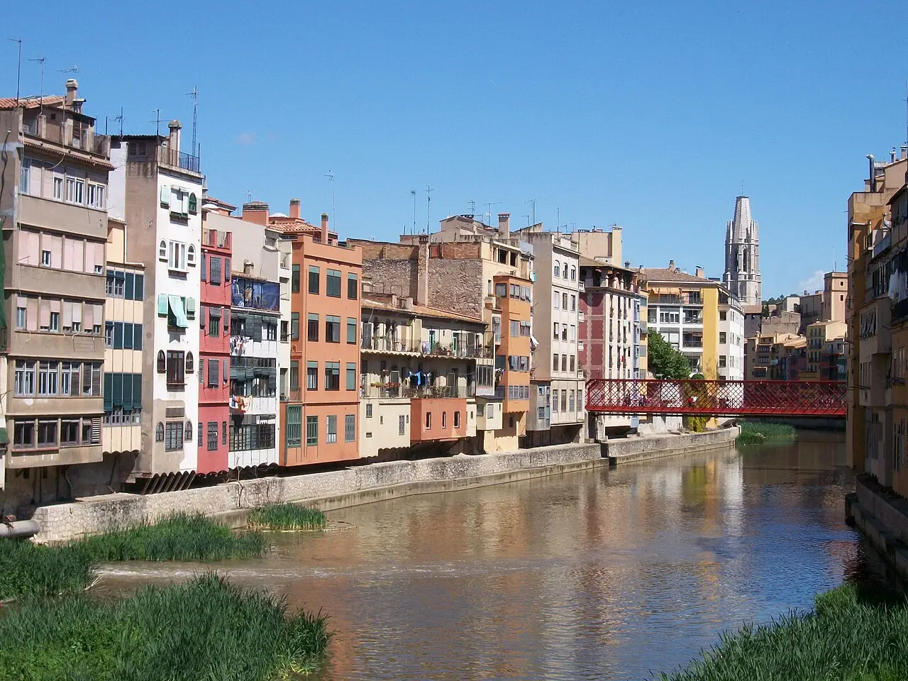 Casas coloridas à margem do rio Onyar em Girona, com a ponte Eiffel de estrutura vermelha e uma torre de igreja ao fundo.