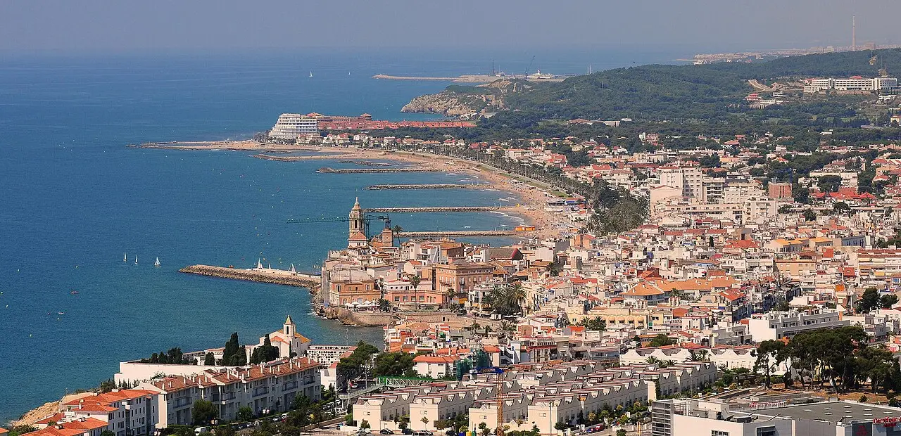 Vista aérea de Sitges, com a igreja junto ao mar, casas de telhado vermelho, a faixa de praia com espigões e o Mediterrâneo à esquerda.