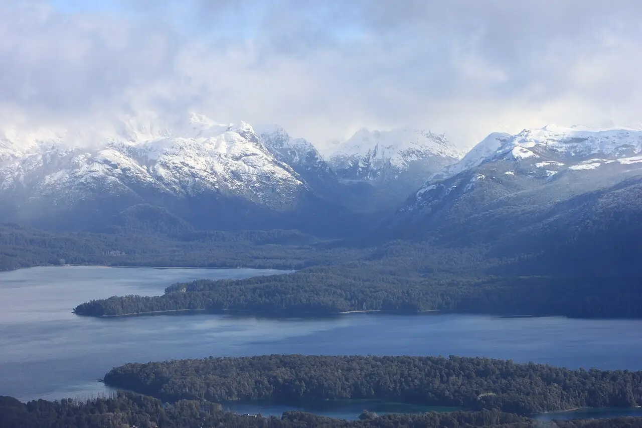 Vista do alto sobre um lago azul-escuro com penínsulas cobertas de bosque e montanhas nevadas com nuvens ao fundo.