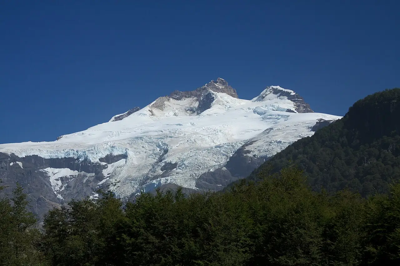 Cume nevado do Cerro Tronador com geleiras descendo pela encosta, contra um céu azul profundo e com floresta em primeiro plano.