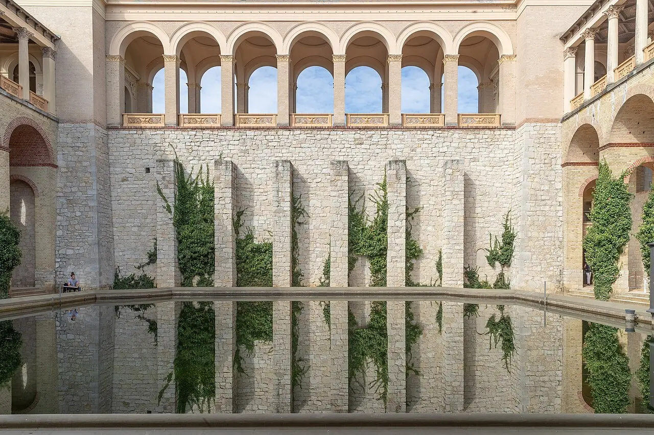 Pátio do Belvedere do Pfingstberg em Potsdam, com uma galeria de arcos no alto, muro de pedra clara com trepadeiras e o reflexo num espelho d’água.